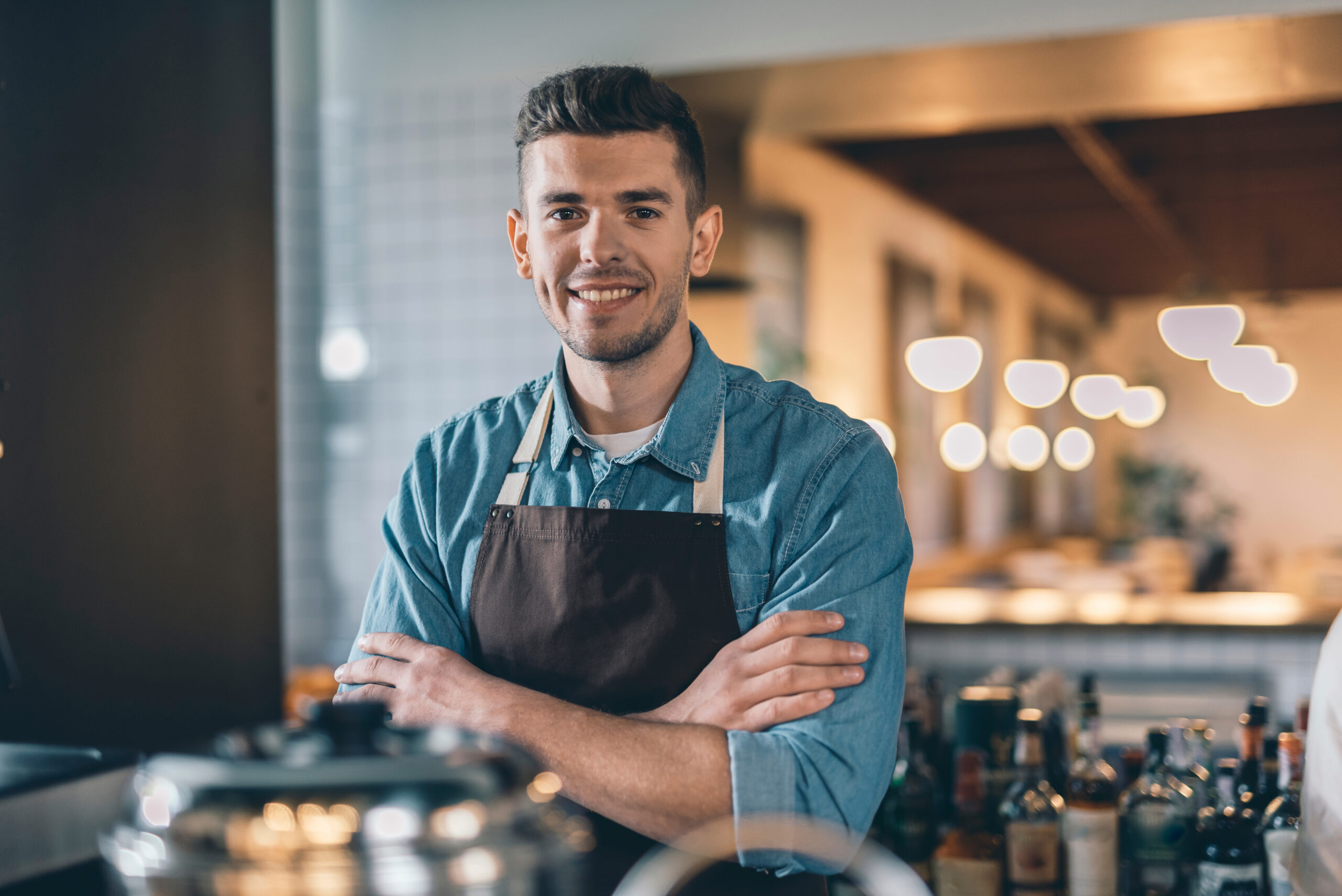 Smiling man in a denim shirt and brown apron stands confidently with crossed arms in a modern café, showcasing a welcoming atmosphere.