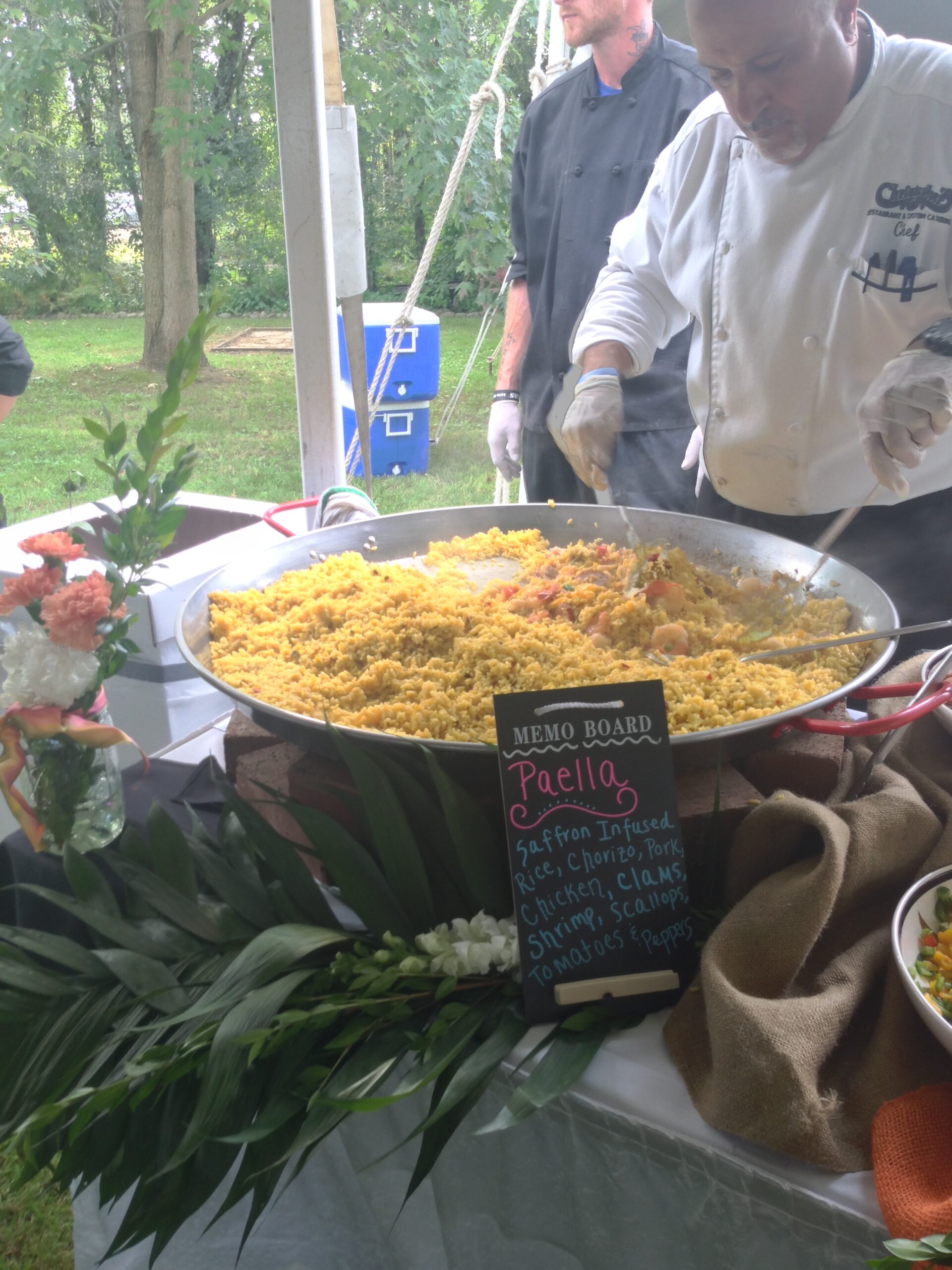 A chef stirs a large paella dish featuring saffron-infused rice, seafood, and meats, with a colorful menu board listing ingredients nearby.