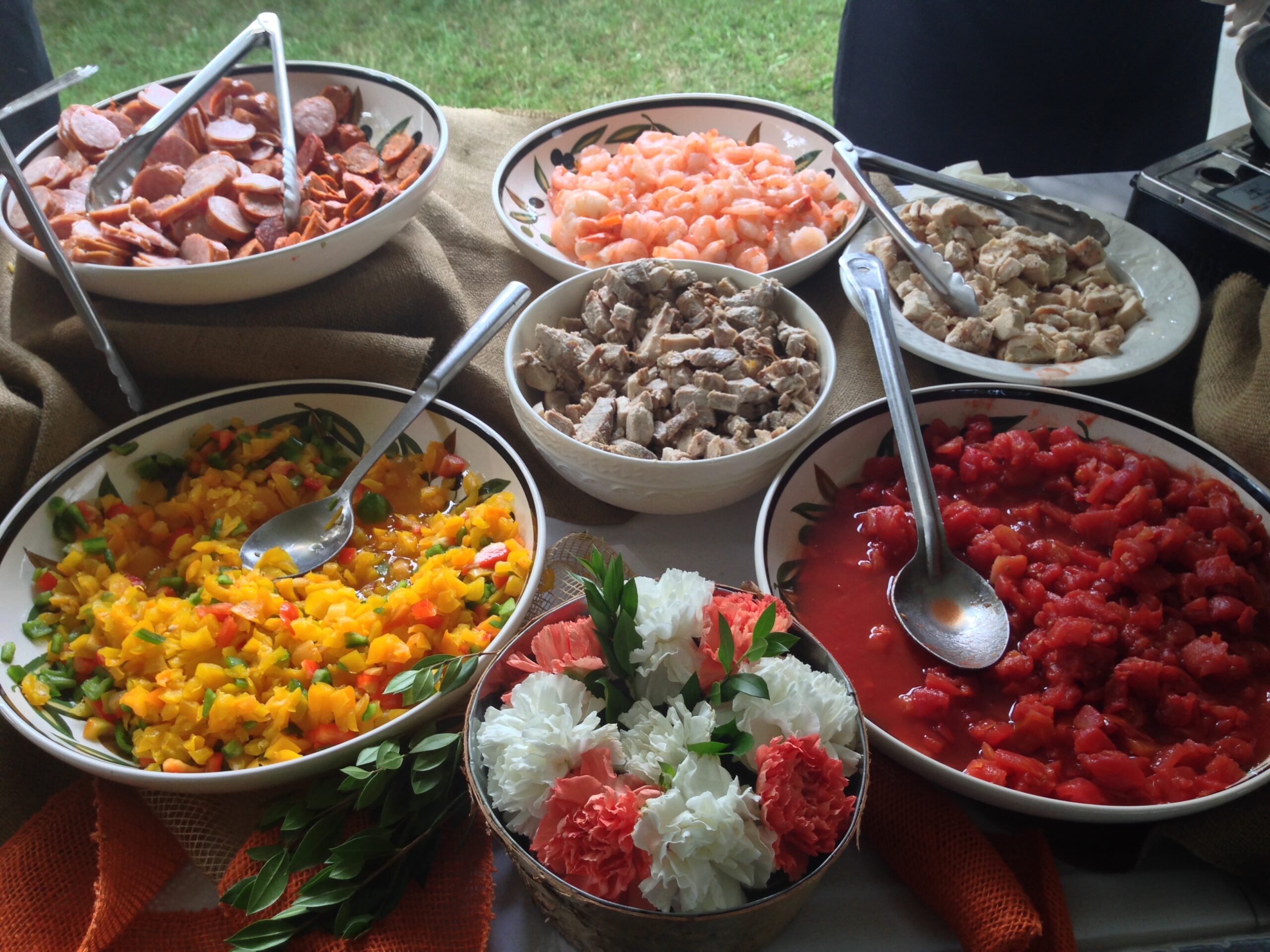 A colorful spread of various dishes, including sliced meats, shrimp, and diced vegetables, arranged in bowls on a table. Fresh flowers accent the display.
