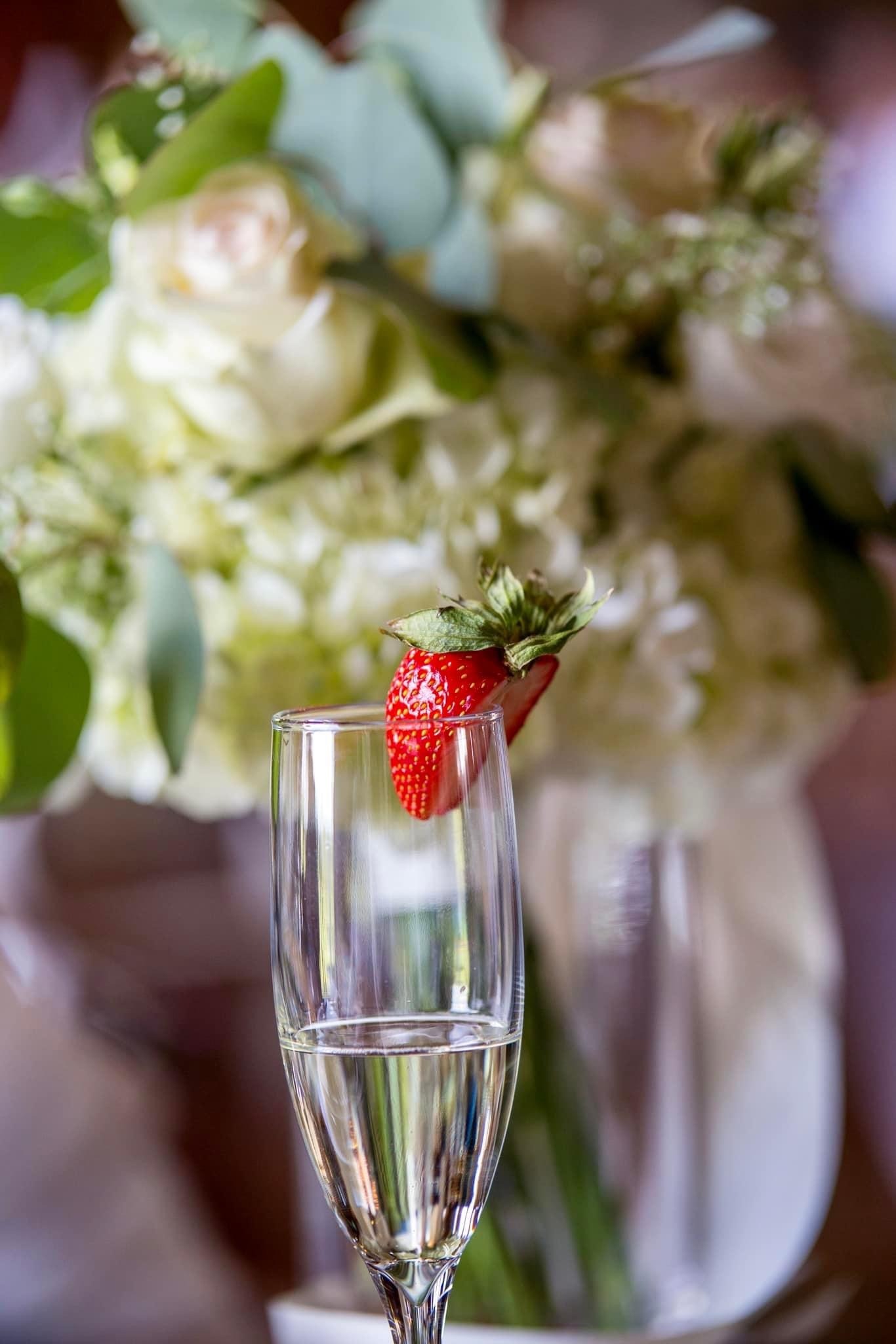 A champagne flute garnished with a strawberry sits in front of a bouquet of white flowers, evoking an elegant celebration atmosphere.