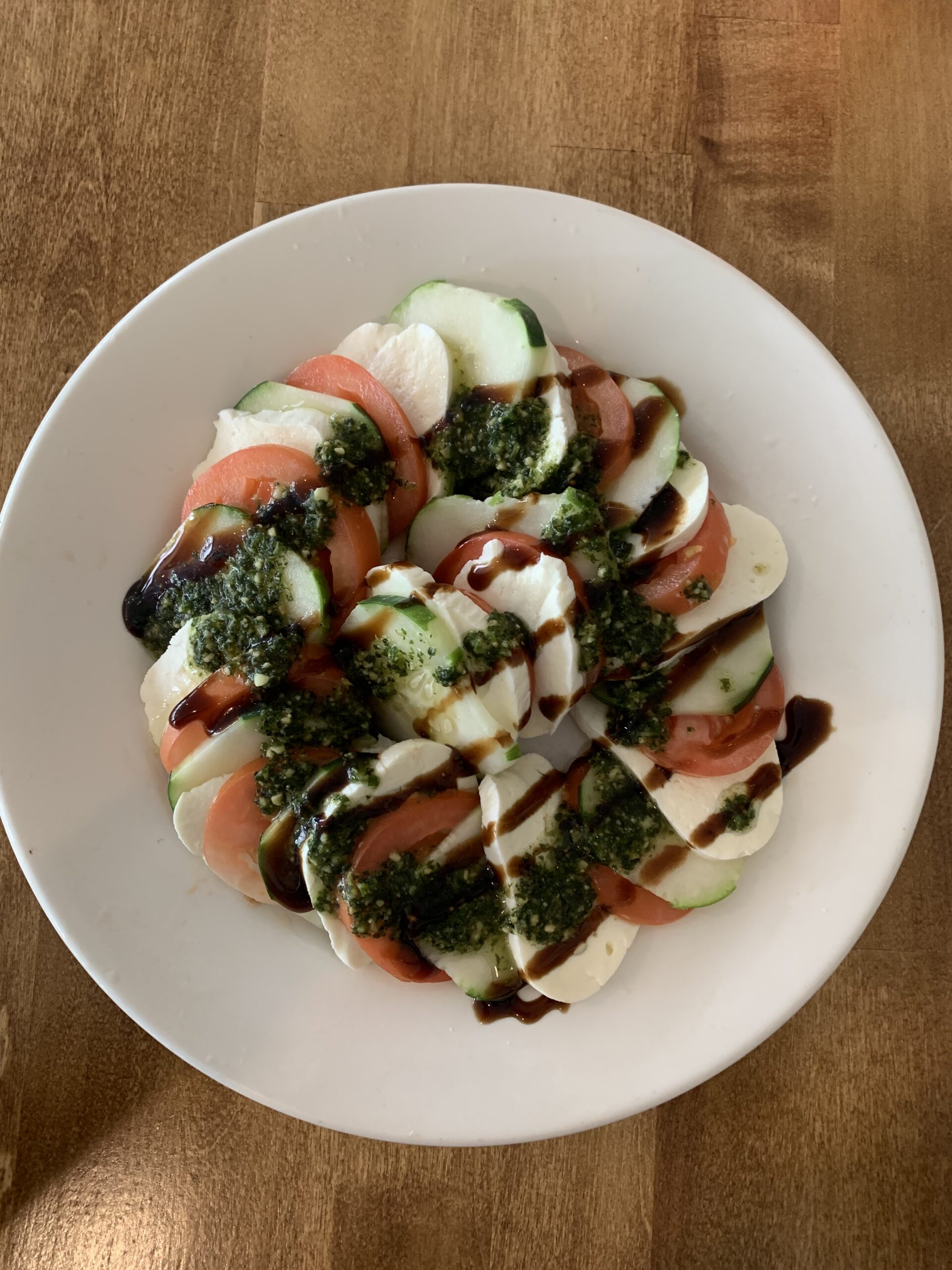 Caprese salad featuring layers of fresh mozzarella, tomatoes, and cucumbers drizzled with pesto and balsamic glaze, served on a white plate.