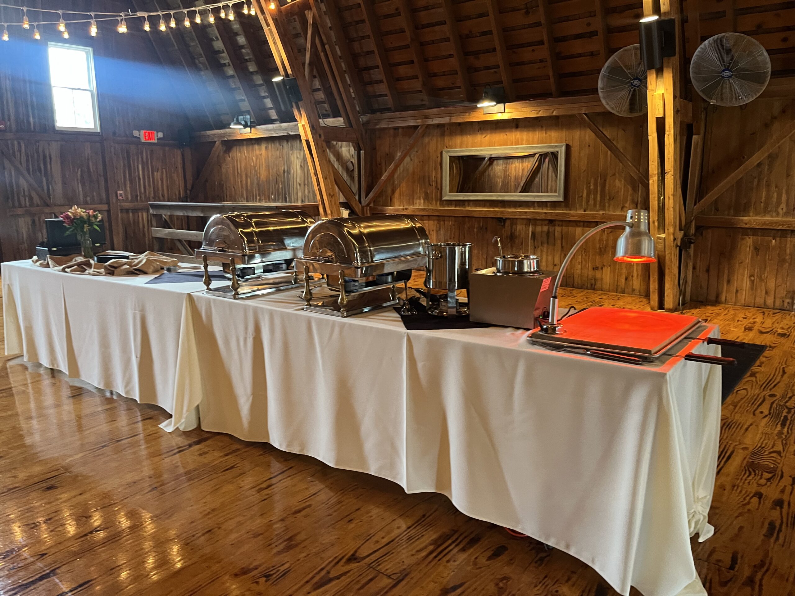 Buffet setup in a rustic barn features silver chafing dishes, a warming lamp, and floral decor on a white tablecloth, ready for a meal service.