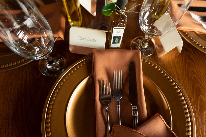 Elegant dining setting featuring place card for "Claire Lander," polished cutlery, wine glass, and decorative olive oil bottle on a wooden table.