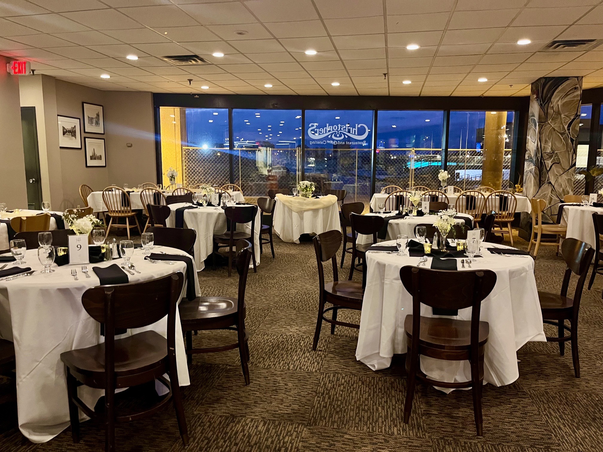 Elegant restaurant interior with round tables set for dining. Soft lighting highlights the decor and window showcasing an evening view.