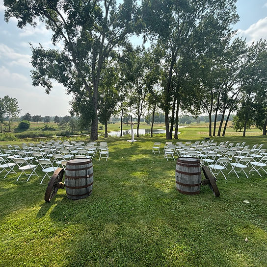 Outdoor wedding ceremony setup featuring rows of white chairs, decorative barrels, and a backdrop of trees and a serene pond.