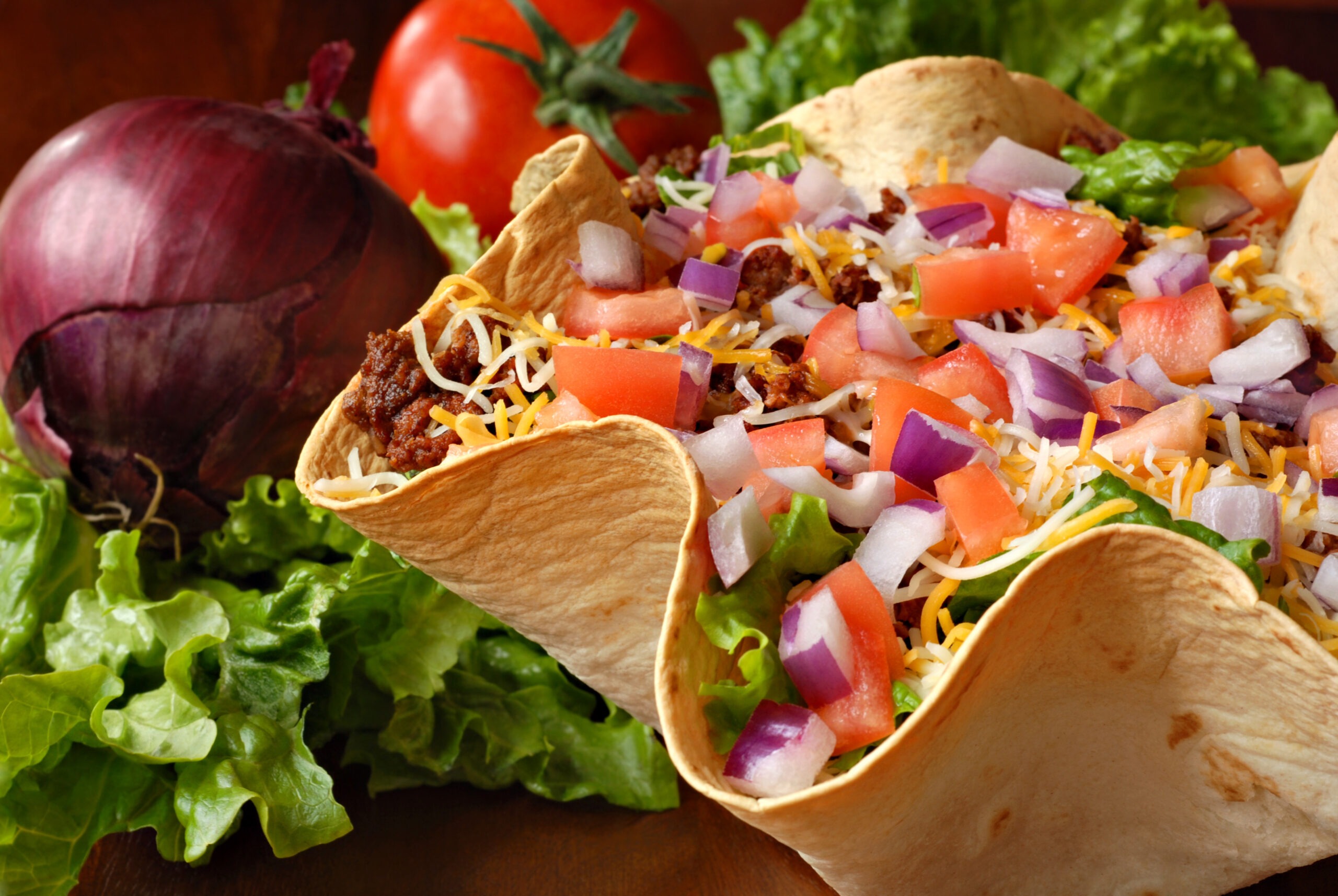 Taco salad in freshly baked flour tortilla bowl (seasoned ground beef, lettuce, onions, tomatoes, and shredded cheese) with tomato, onion, and leaf lettuce in background. Macro with shallow dof.